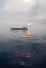 Obraz premium A large cargo ship is anchored on the exceptionally calm waters of Ternate, Indonesia, at dawn or dusk. A peaceful and minimalist scene with the sea reflecting the soft, cloudy sky.