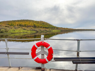 View from a ferry deck showing a red lifebuoy in the foreground, overlooking a calm river with a...