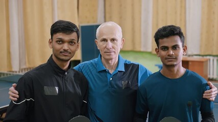 Coach stands proudly with two young players holding table tennis rackets during indoor photoshoot, symbolizing unity, mentorship, teamwork, training, confidence, and dedication - Powered by Adobe