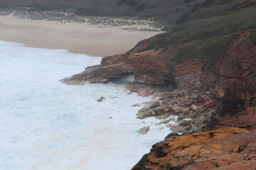 Rocky coastline of Nazare, Portugal with waves crashing against cliffs and sandy beach in the background, dramatic seascape with people on the viewpoint.