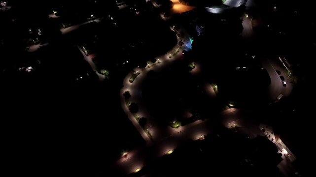 Aerial top down tracking shot of car with xenon headlights on street of quiet neighborhood. Orange lights of streetlamp shining on asphalt. American suburbia at night.