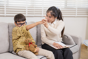 Young child sitting on sofa hugging teddy bear tightly, avoiding eye contact with therapist during...