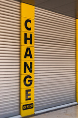 A change office is seen with bold letters on the sign, while its shutters are closed. The bright yellow and black colors draw attention to the establishment in a busy urban setting