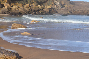 Rocky seashore with gentle waves rolling onto sandy beach under sunlight, creating a peaceful coastal landscape with ocean spray and natural textures.