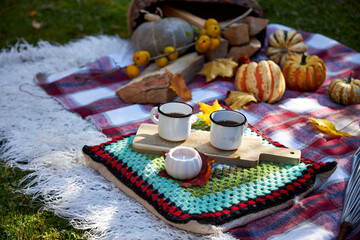 Cozy autumn picnic in the garden with pumpkins, colorful leaves, hot tea in enamel mugs, candlelight and warm blanket. Rustic fall atmosphere, relaxation and seasonal outdoor decoration