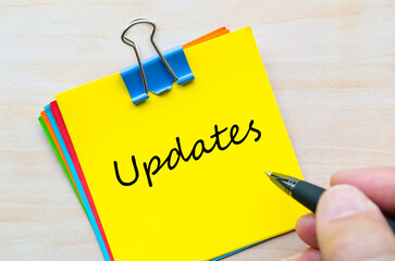 Updates handwritten text on yellow notepad clipped with silver pin on light wooden desk.