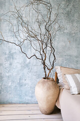 A large ceramic vase with dry branches stands against a textured blue wall. The scene is minimalistic and modern, emphasizing natural elements in interior design.