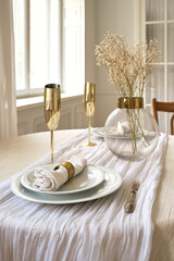 Elegant dining table set with white tablecloth, gold-accented glasses, and decorative flowers in a glass vase. Soft natural light enhances the ambiance.