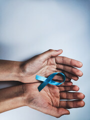 Fototapeta premium ​Hands gently holding the light blue awareness ribbon, a symbol for conditions like Prostate Cancer and Thyroid Disease. Health and support concept on a simple background.