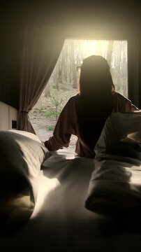 Woman waking up in bed, looking out window at the morning sunlight in a forest setting.