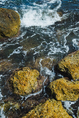 Waves crashing over rocks along a vibrant coastal shore at midday under a clear blue sky in a serene seaside setting