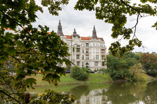 Grand neoclassical mansion with ornate domed towers and red roofs surrounded by green park and river with natural tree frame on overcast day. Historic architecture, luxury estates, European heritage.