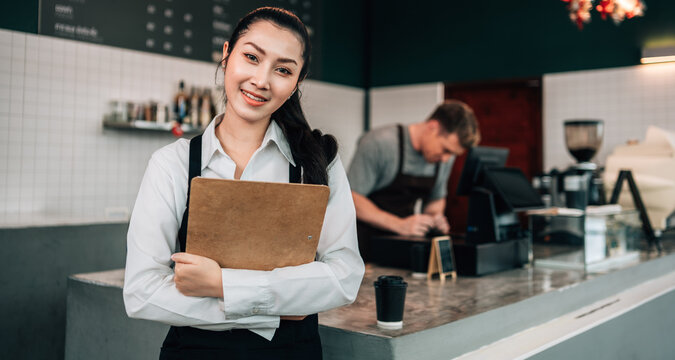 A smiling female cafe owner or manager stands behind the counter, holding a clipboard and looking directly at the camera. A male barista is working in the background.