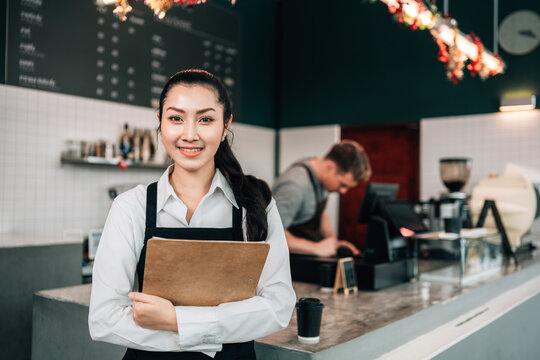 A smiling female cafe owner or manager stands behind the counter, holding a clipboard and looking directly at the camera. A male barista is working in the background.