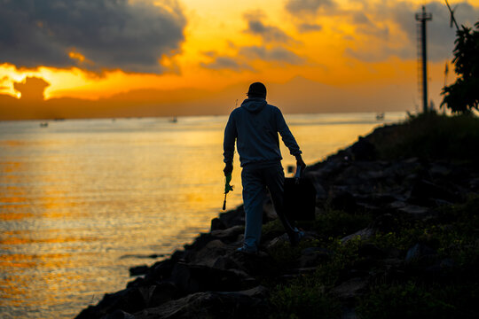 Man with fishing rod walking along rocky shore at sunrise, silhouette against a vibrant golden sky. - Powered by Adobe