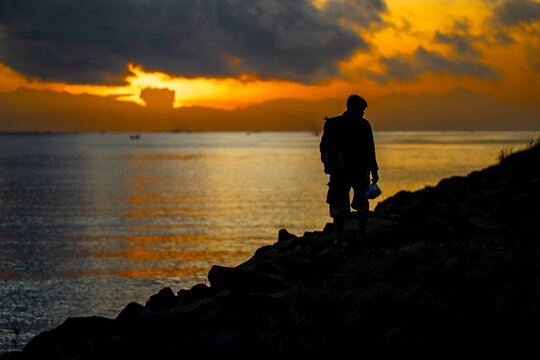 Man with fishing rod walking along rocky shore at sunrise, silhouette against a vibrant golden sky.