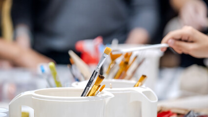 A close-up shot of paintbrushes in the foreground, with two women artists working on handmade products in the background of an art studio