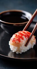 A single piece of sushi, topped with vibrant red seafood, is delicately lifted by chopsticks from a black plate, alongside a small bowl of soy sauce