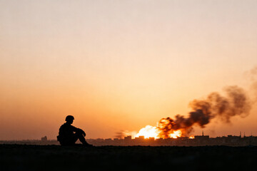 Silhouette of a person sitting on a hill at sunset with fire and smoke in the distance