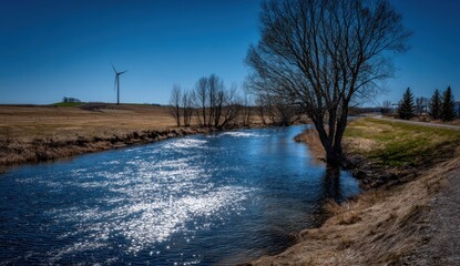 A serene rural landscape showcasing a sparkling river, bare trees, and a wind turbine against a vibrant blue sky.