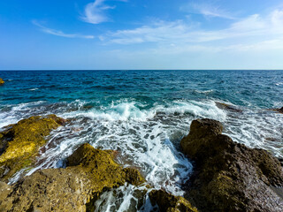 Waves crash against rugged rocks along the serene coastline under a bright blue sky on a sunny day by the ocean