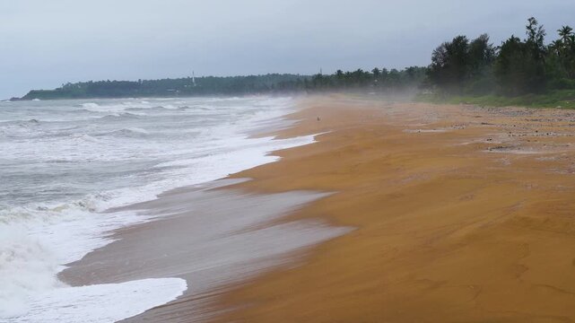 Rough waves on the sandy coast of Bekal, cochin, kerala, Mangalore during a monsoon storm with fog and spray rising in the distance covering the trees and buildings
