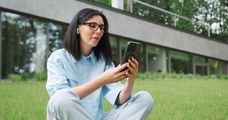 Cheerful young Caucasian woman in casual clothes sitting cross-legged on the grass in a city park, using her smartphone for a video call. Concept of wireless communication, remote work - Powered by Adobe