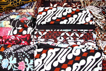 Obraz premium Colorful batik fabrics stacked high in market stall display intricate patterns and vibrant colors, showcasing traditional craftsmanship and cultural richness.