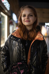 Young woman with long brown hair wearing a stylish black puffer jacket stands confidently in an urban setting, showcasing modern fashion and vibrant city life
