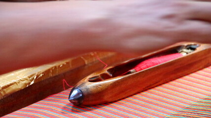 Wooden shuttle carrying red thread rests on colorful woven fabric in a textile mill, showcasing traditional craftsmanship and vibrant colors.