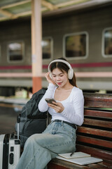 Asian woman sitting at a railroad station with backpack, using mobile phone and laptop, waiting for train, enjoying travel lifestyle.