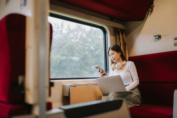 Fototapeta premium Smiling young woman traveler with suitcase and headphones around her neck holding a smartphone at the train station.