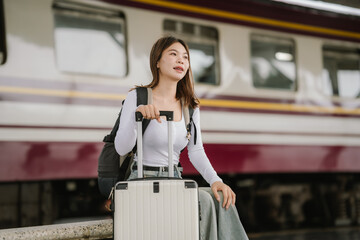Smiling young woman traveler with suitcase and headphones around her neck holding a smartphone at the train station.