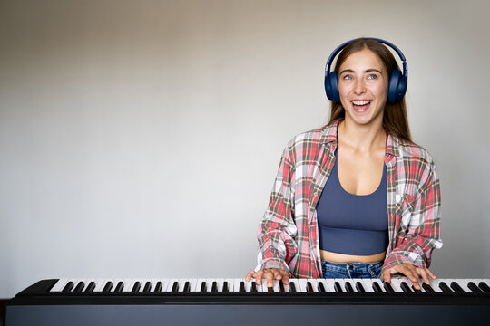 Portrait of young woman smiling while playing digital piano with headphones indoors, music education practice