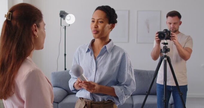 Female journalist with microphone interviewing young woman while cameraman filming on background. Smiling reporter listening to guest and collecting information while conducting media interview.
