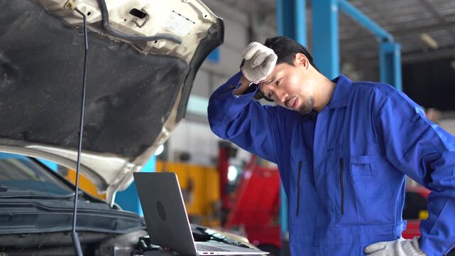 A frustrated Asian mechanic wipes sweat while facing a complex computer diagnostics issue with an open-hood vehicle. Japanese mechanic man in blue uniform Stressed car mechanic in repair garage
