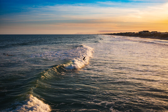 A lone surfer riding a small wave in as seen from a pier near sunset