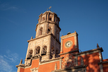 A tall, red building with a clock tower and a cross on top. Historic center of Queretaro City, decorations and traditions to celebrate Mexico's Independence Day, colonial architecture, alleys and faca