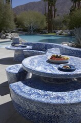 Outdoor patio dining set with a blue and white mosaic pattern, featuring a round table and benches, near a swimming pool, on a sunny day.