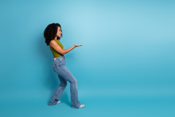 Young woman with curly hair posing casually in front of blue background, gesturing with hands and smiling joyfully