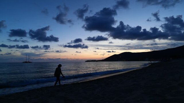 Ogliastro Marina, Italy. Blue hour view of Ogliastro Marina Bay, Italy: a lone person walks on Baia Arena beach as waves roll in; a 2-masted sailing ship anchors off Punta Ogliastro after sunset.