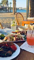 The image shows a table with seafood dishes, salad, bread, and drinks on a terrace overlooking the sea. The setting is perfect for a relaxing meal with a view of the beach and palm trees.