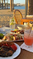The image shows a table with seafood dishes, salad, bread, and drinks on a terrace overlooking the sea. The setting is perfect for a relaxing meal with a view of the beach and palm trees.