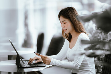 Young woman holding credit card and smartphone, making online payment while using laptop in modern cafe.