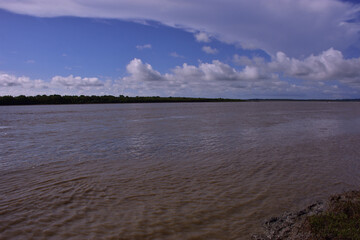 Scenic wide-angle view of the Bakkhali River in Cox’s Bazar, Bangladesh, with flowing water under a dramatic blue sky filled with white clouds. A natural landscape of river and sky.