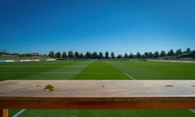 Empty wooden bench, overlooking a soccer field under a clear blue sky