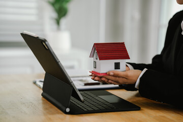 Businesswoman writing notes with house model, keys, contract, and calculator on desk, symbolizing property investment, real estate planning, and financial agreement.