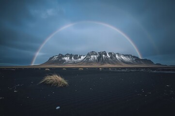 A majestic mountain range with snow-capped peaks is framed by a vibrant rainbow arching across a cloudy sky, while the foreground features a dark, textured landscape.