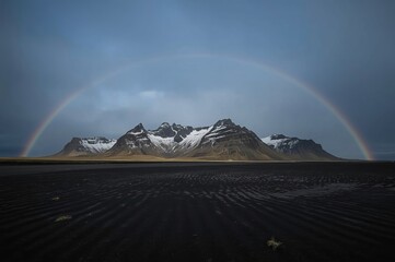 A majestic mountain range with snow-capped peaks is framed by a vibrant rainbow arching across a cloudy sky, while the foreground features a dark, textured landscape.