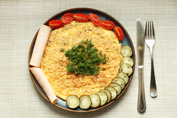 An oatmeal omelet sits on a blue plate alongside lightly salted cucumbers, tomatoes, and ham. Top view. Closeup.